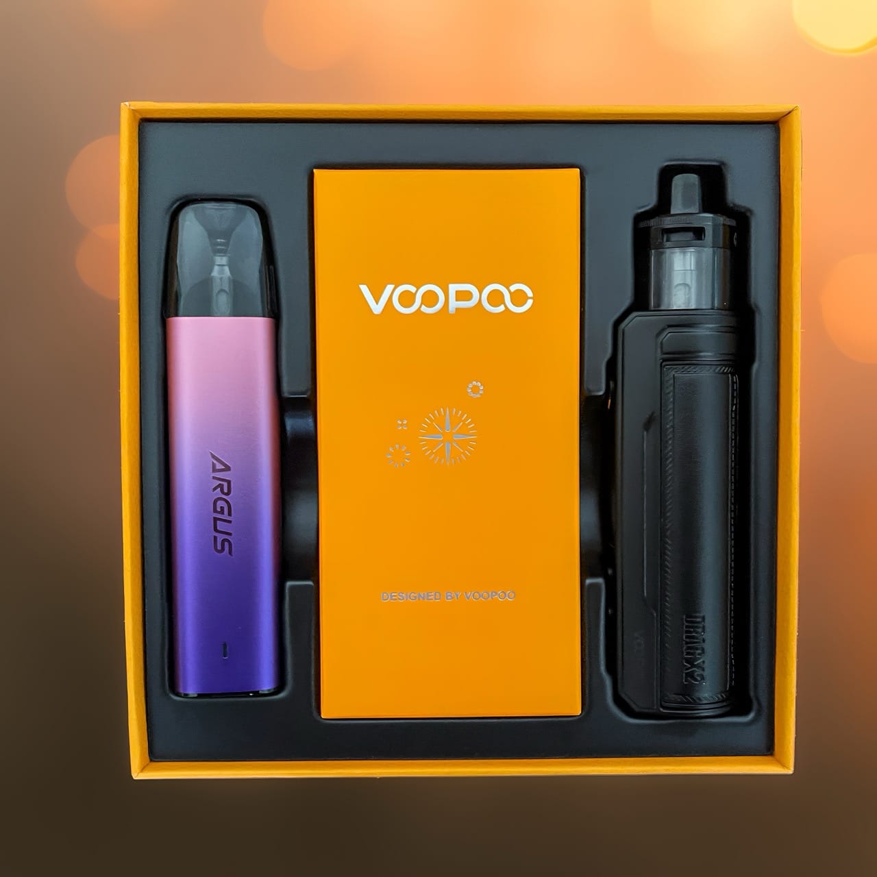 Vue plongeante sur le coffret Bonnes Vacances Voopoo ouvert, présentant l'ARGUS G2 Mini violet et le DRAG X2 noir disposés de part et d'autre d'un livret orange